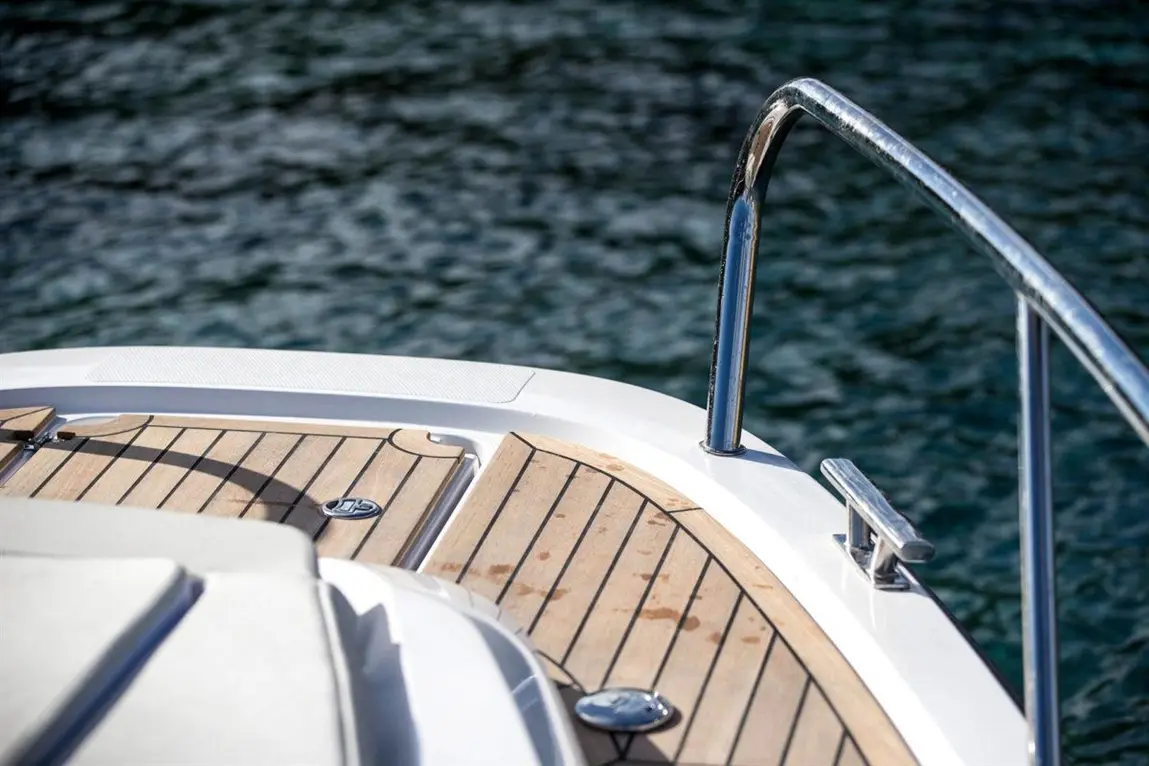 Close-up of a boat's deck showing polished wood, stainless steel railing, and glistening water in the background.